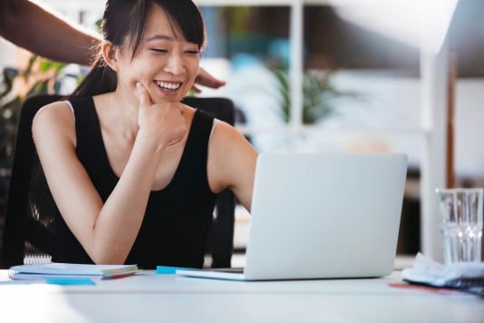 Shot of young smiling woman sitting at her desk and working on laptop. Asian female executive using laptop computer.