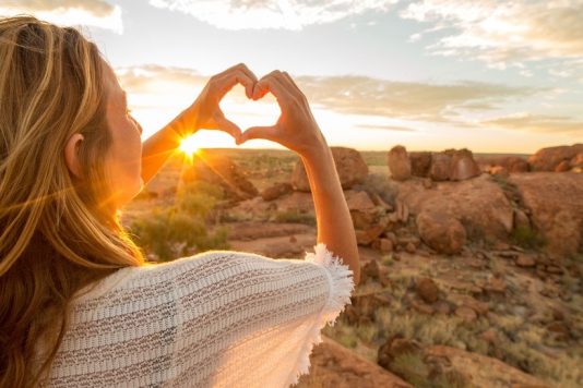 Young woman at the devil's Marbles makes a heart shape finger frame to the spectacular landscape at sunrise. Devils Marbles Conservation Reserve, Northern Territory, Australia.